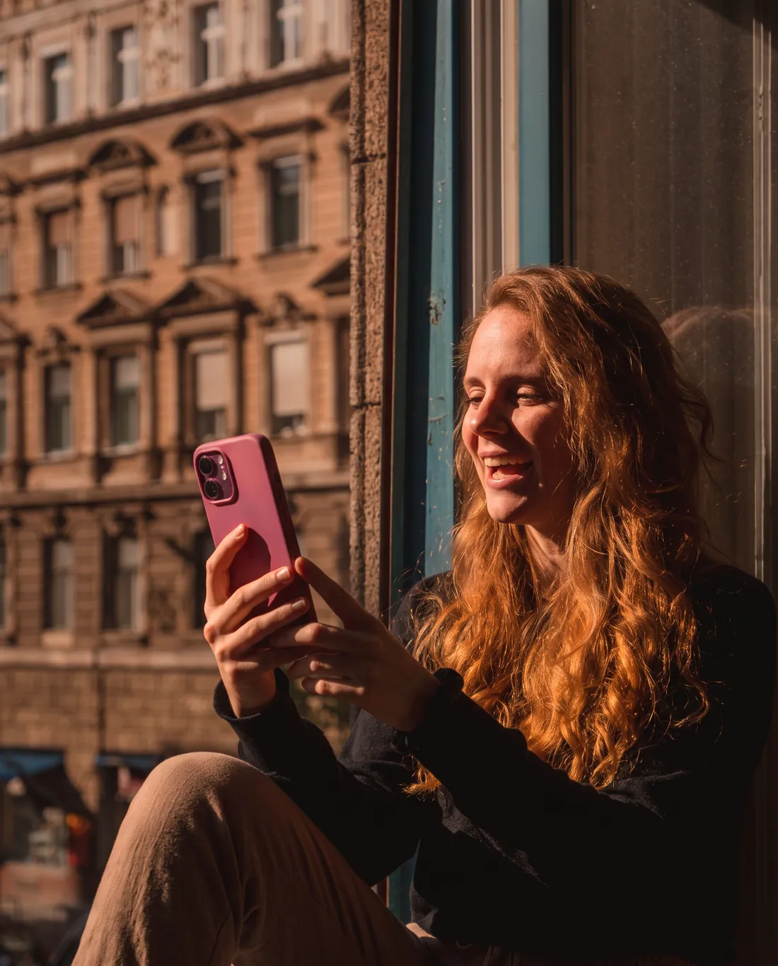 Mulher sorrindo enquanto usa o celular perto da janela